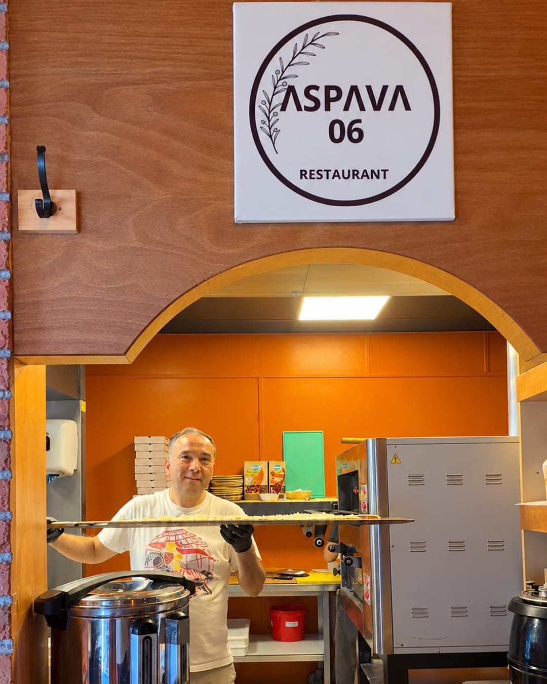 De meester met een pideschep achter de keukenboog, erboven het ASPAVA 06-bord (glimlach bewerkt met AI)