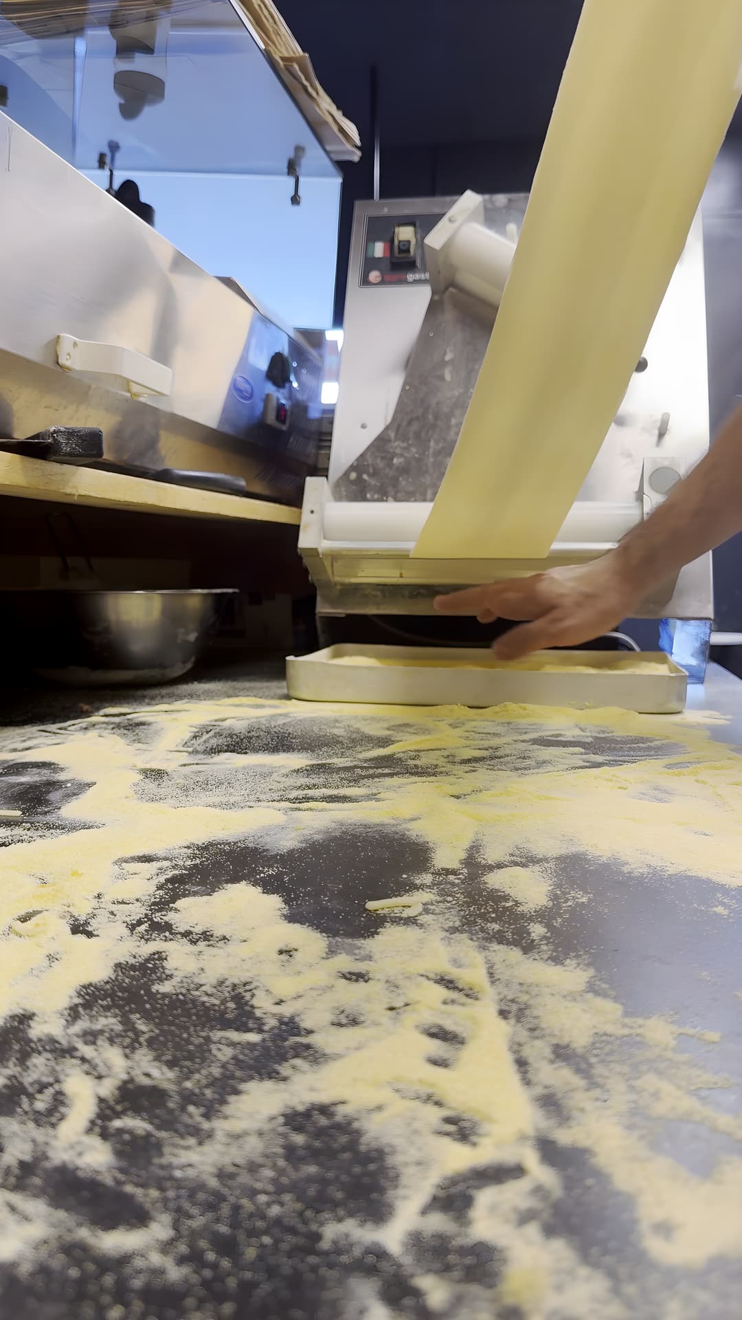 Een strook deeg komt uit een deegmachine, met meel op het werkblad en een hand eronder